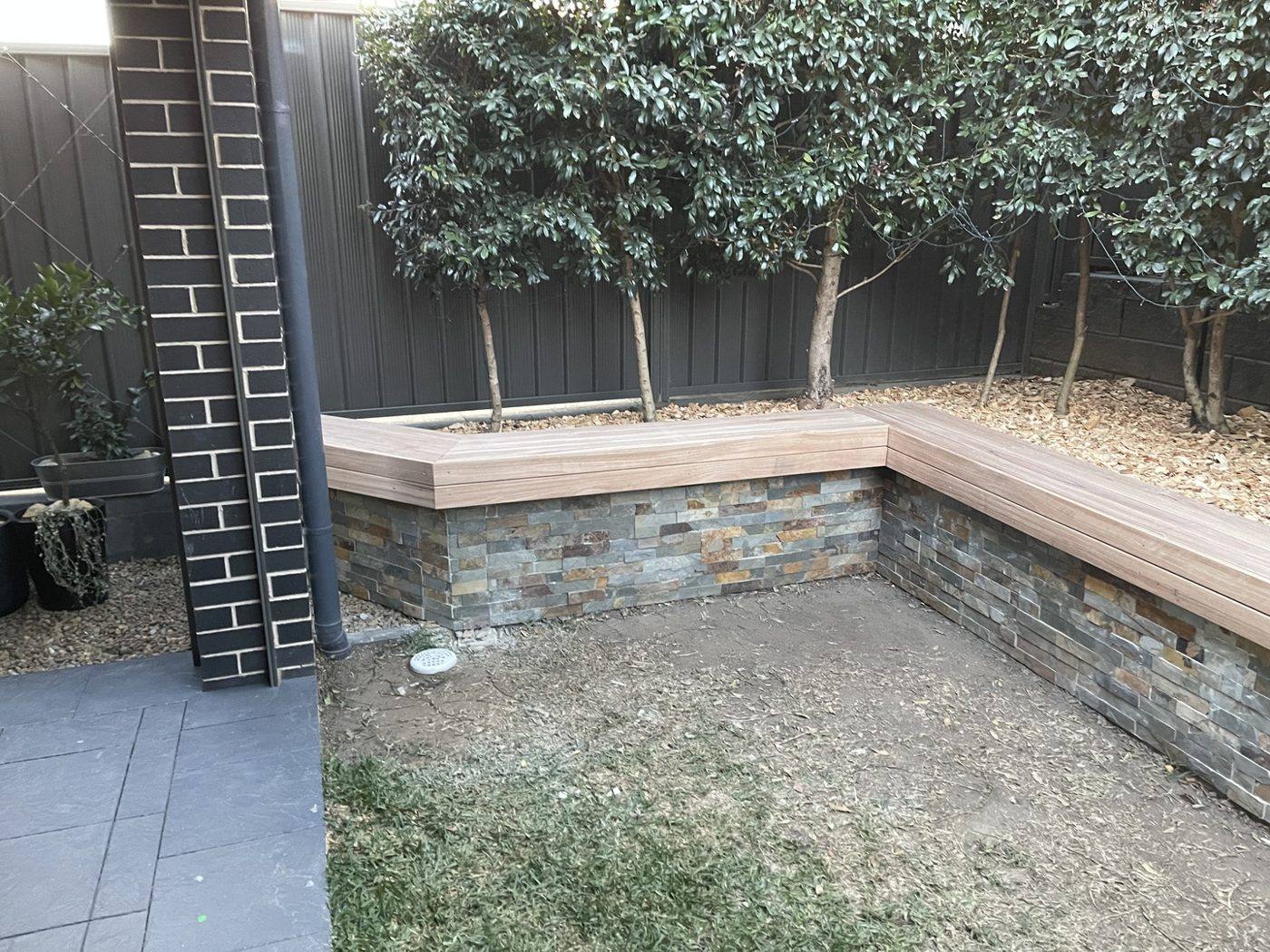 Finished pale timber bench seat wrapping the corner of a stone-clad planter wall, courtyard by Loel Homes