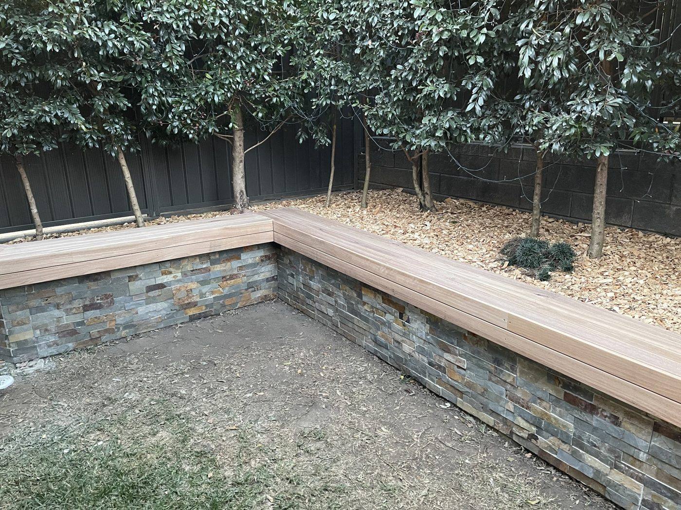 Finished L-shaped timber bench seat along two stone planter walls in a courtyard by Loel Homes