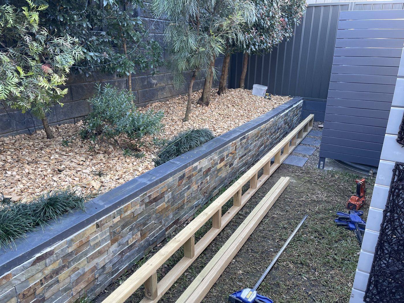 Timber bench rails being laid out along the stone-clad planter wall in a courtyard by Loel Homes