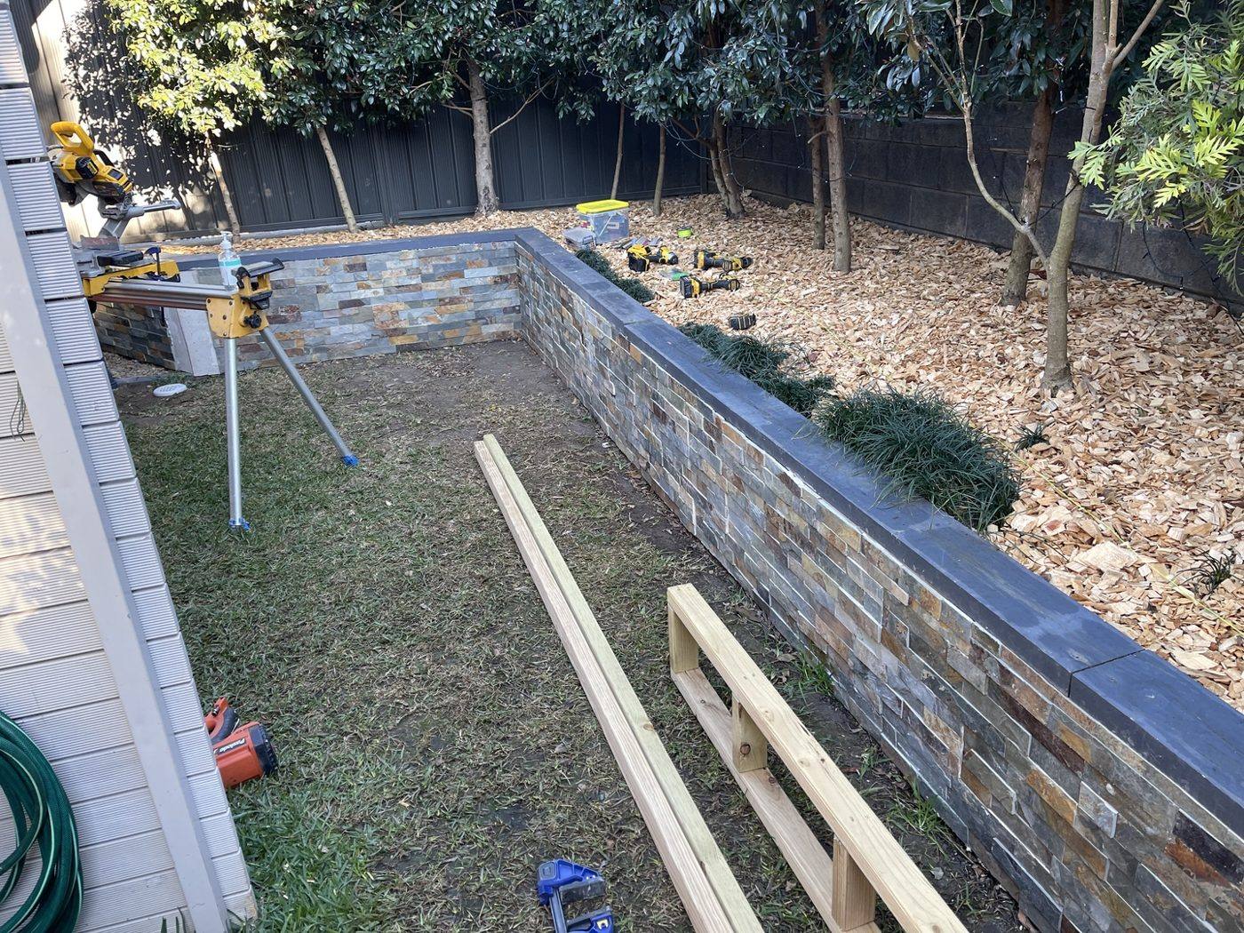 Finished stone-clad planter wall with a drop saw set up and timber bearers laid out ready to build the bench, courtyard by Loel Homes