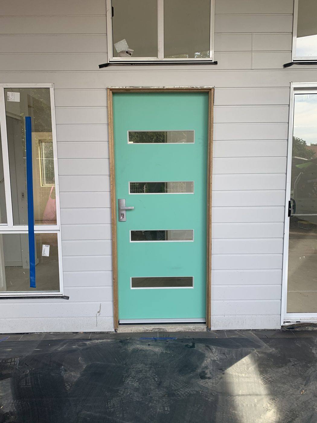 Teal four-slot modern front entry door with a lever handle set on a white weatherboard home, front view by Loel Homes