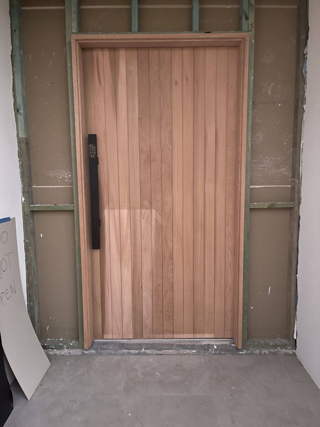 Oversized vertical-batten timber front entry door hung in a rendered opening with a long pull handle by Loel Homes