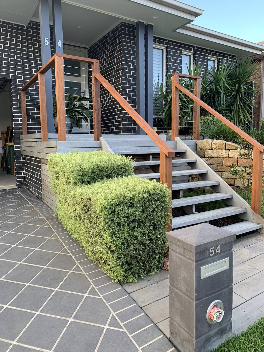 Finished front entry stairs with spotted-gum timber posts, stainless wire balustrade and rendered landing on a dark-brick home by Loel Homes