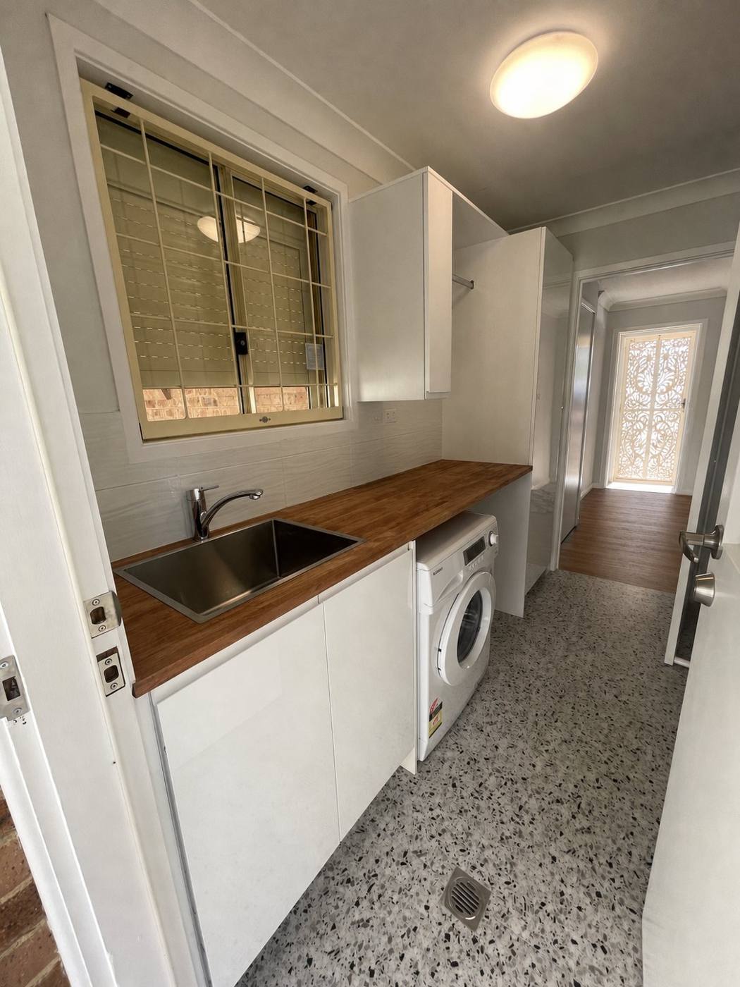 Finished laundry after renovation with stainless sink, timber benchtop, white gloss cabinets, front-loader washer, overhead cabinet with hanging rail and terrazzo floor, Quakers Hill by Loel Homes