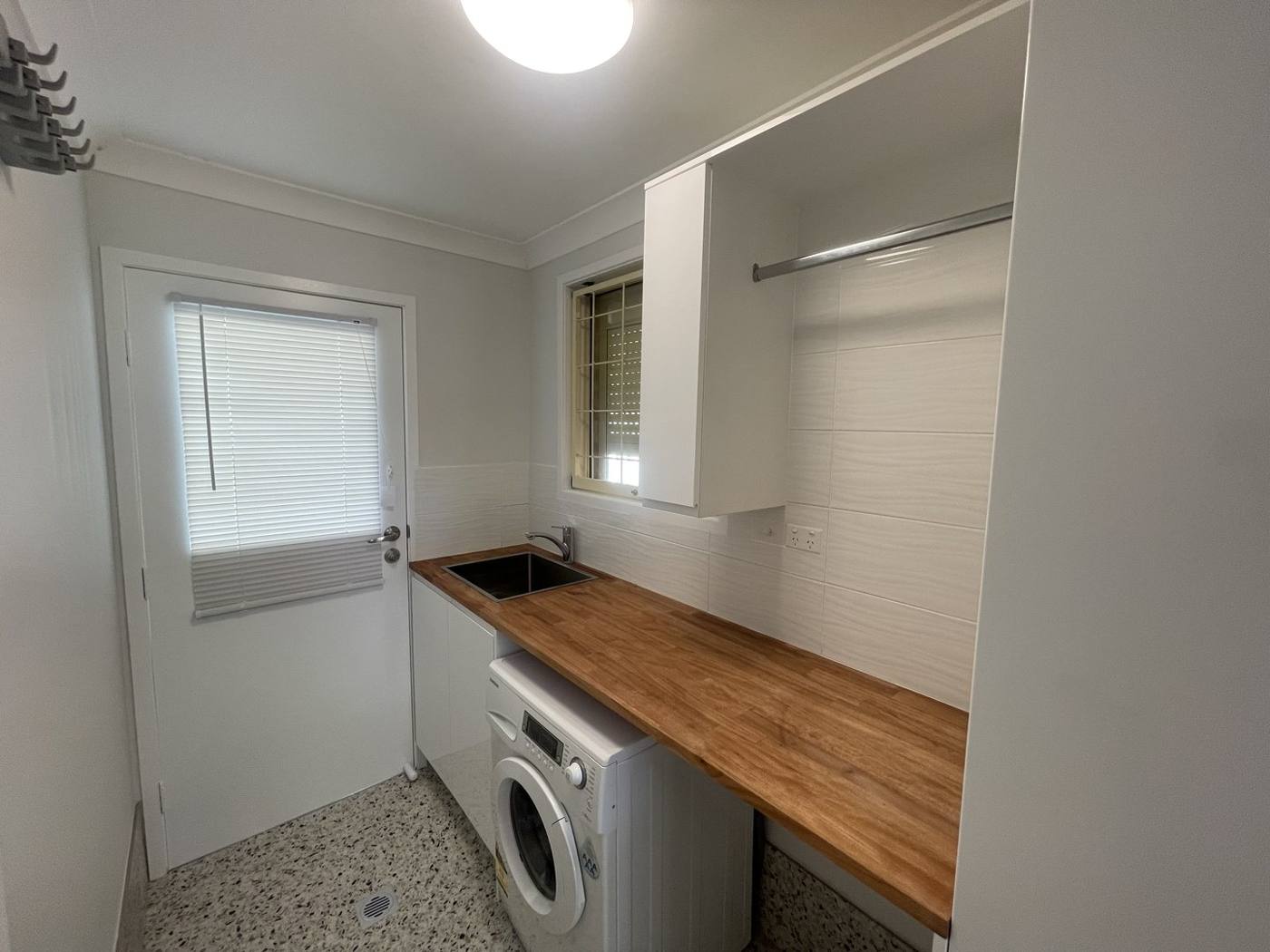 Finished laundry after renovation with timber benchtop, stainless sink, front-loader washer, overhead cabinet with rail, white tiled splashback and external door, Quakers Hill by Loel Homes