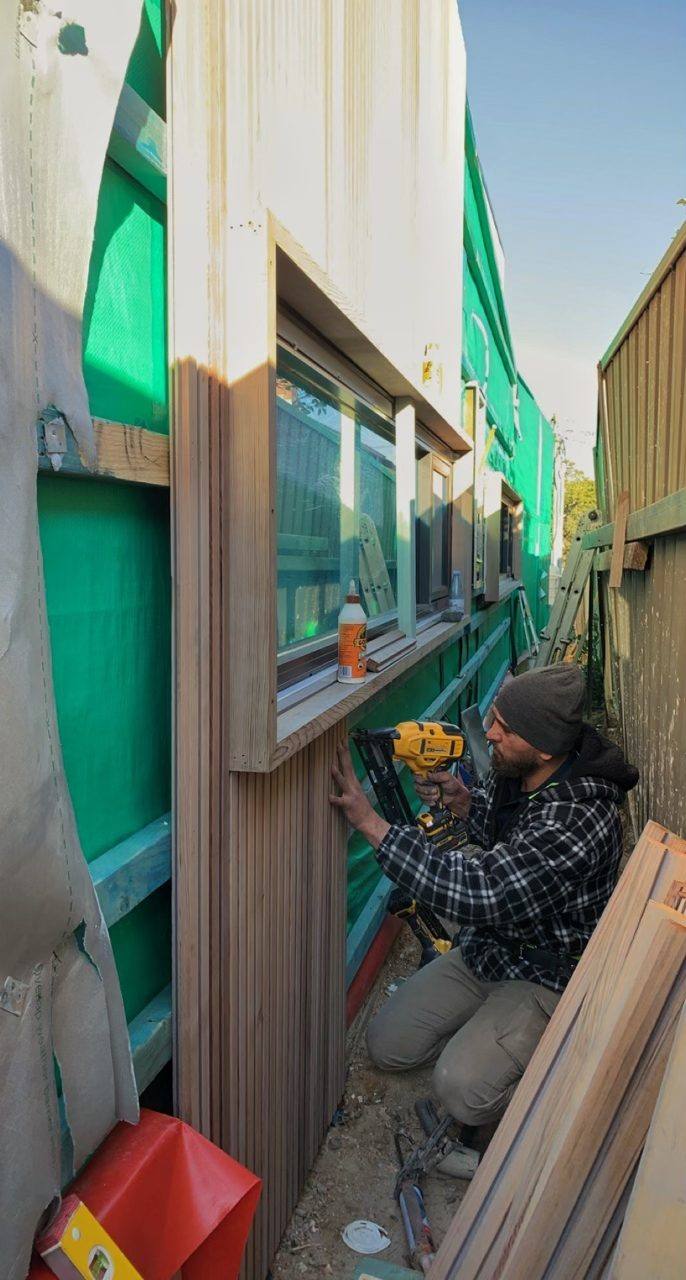 Tradesman nail-gunning pale timber cladding boards around a window over green wall wrap, duplex by Loel Homes